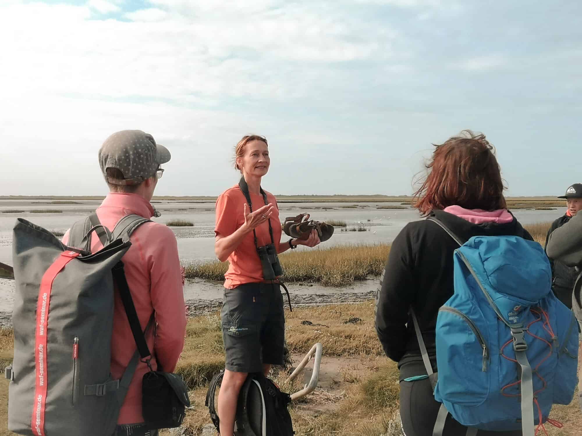 Group of birdwatchers with backpacks engaged in outdoor activity by a coastal marshland, a woman in orange addressing the group with binoculars in hand, enhancing a team-building nature excursion